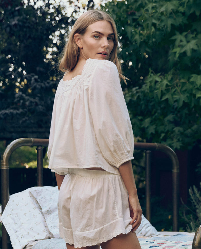 A woman stands outdoors in THE GREAT. CORE SLEEP’s Eyelet Button Sleep Top in True White, its Victorian lace details highlighted as she glances over her shoulder amid greenery and a bed frame.