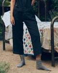 A person stands outdoors beside a metal bed with a colorful quilt, wearing THE GREAT. CORE SLEEP's The Eyelet Easy Sleep Pant in black, paired with a black shirt and gray socks.