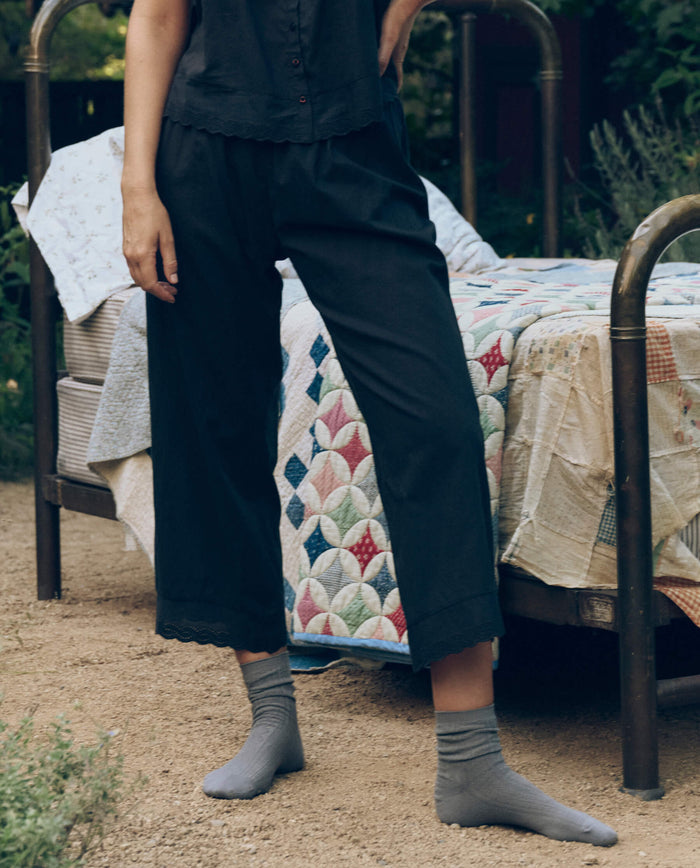 A person stands outdoors beside a metal bed with a colorful quilt, wearing THE GREAT. CORE SLEEP's The Eyelet Easy Sleep Pant in black, paired with a black shirt and gray socks.