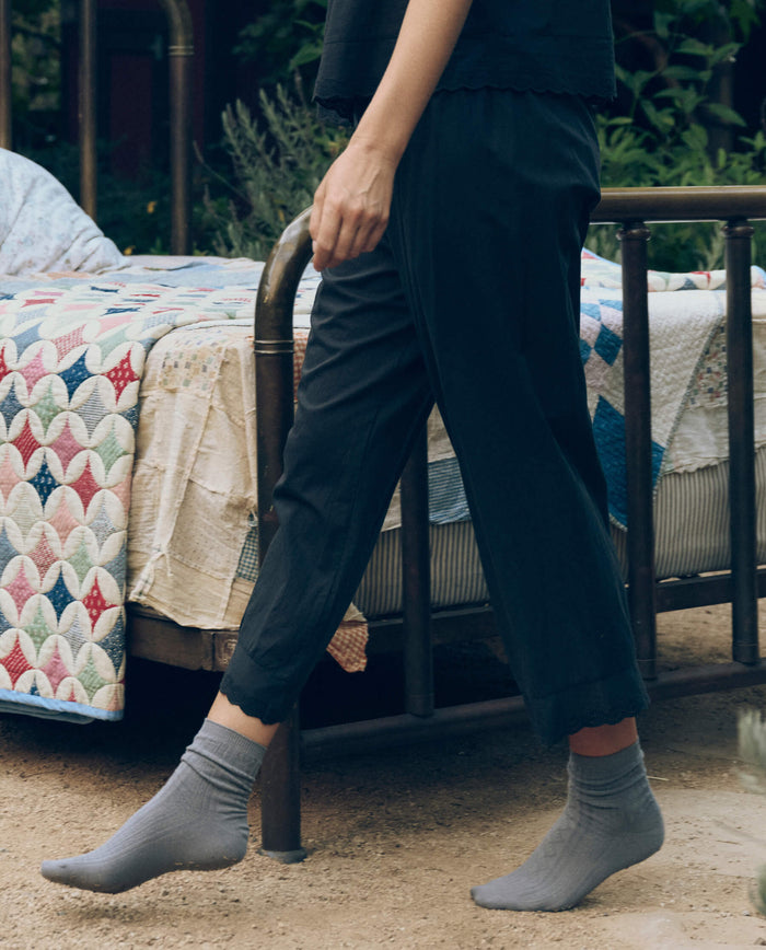 Wearing THE GREAT. CORE SLEEP’s The Eyelet Easy Sleep Pant in black, a person strolls outdoors beside a metal-framed bed topped with a patterned quilt, dressed in Victorian-inspired loungewear.