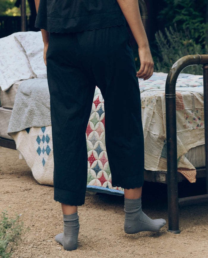 A person wearing THE GREAT. CORE SLEEP The Eyelet Easy Sleep Pant in black and gray socks stands by a vintage-style metal bed with patchwork quilts, outdoors on a dirt surface.