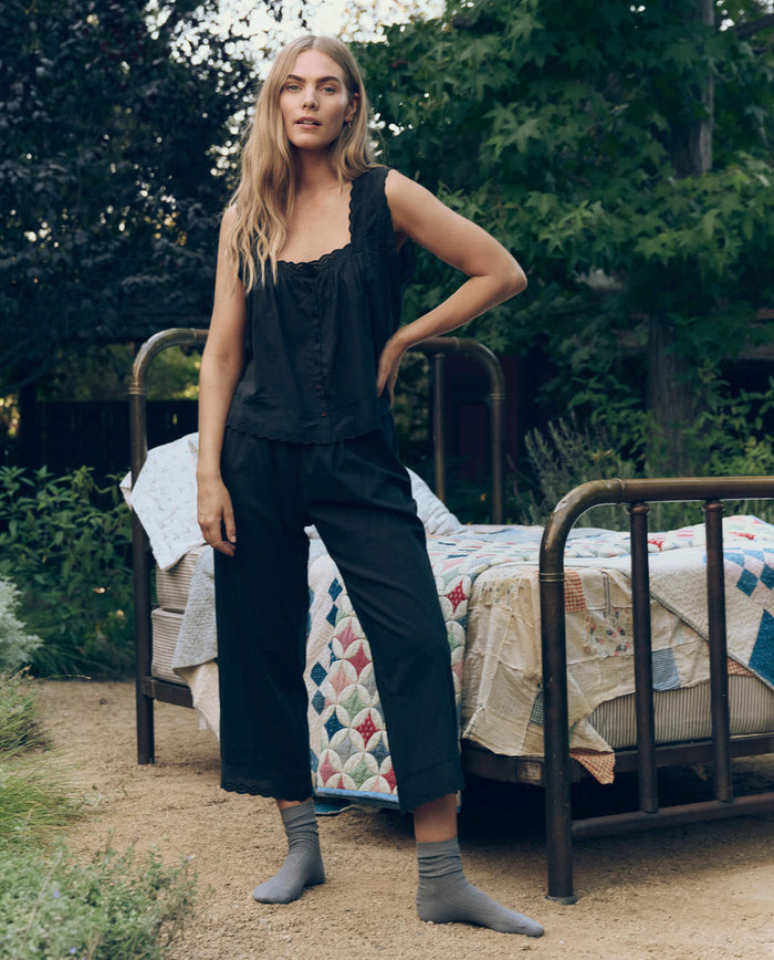 A woman stands outdoors beside a metal bed with a patchwork quilt, wearing THE GREAT. CORE SLEEP’s The Eyelet Easy Sleep Pant in black and gray socks, surrounded by flourishing trees and greenery.