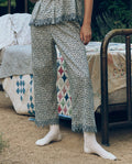 A person wearing The Great. SP26 Sleep Eyelet Easy Sleep Pant in Bluebell Ditsy stands by a metal-frame bed with a colorful quilt outdoors, paired with white socks.