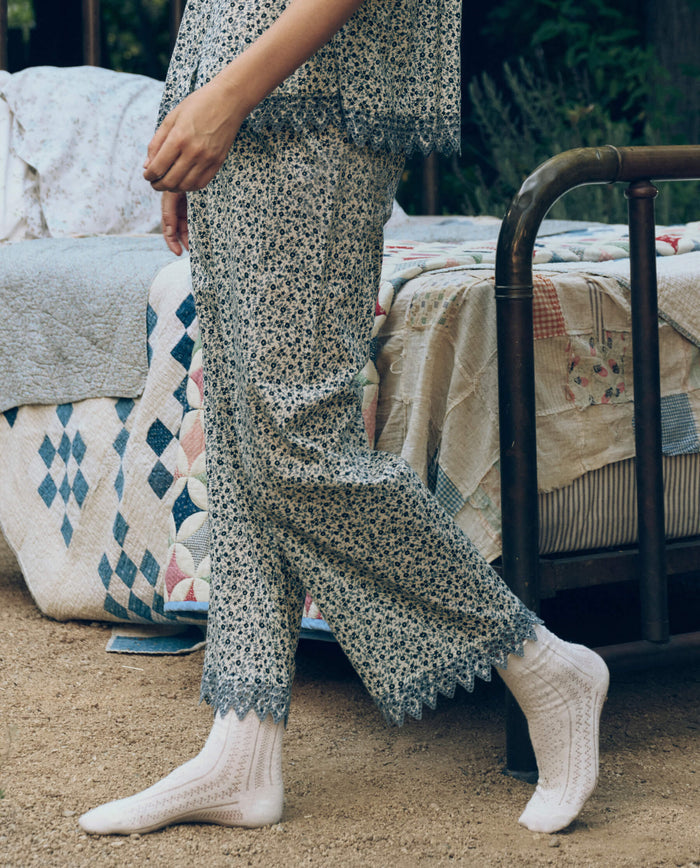 A person in The Great. SP26 Sleep Eyelet Easy Sleep Pant in Bluebell Ditsy and white socks stands beside a vintage metal bed with patchwork quilts outdoors on dirt.