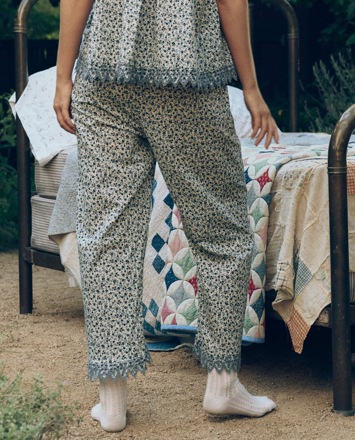 Wearing The Great. SP26 Sleep's Eyelet Easy Sleep Pant in Bluebell Ditsy, a person in patterned pajamas and white socks stands outdoors by a metal bed with a colorful patchwork quilt on dirt.