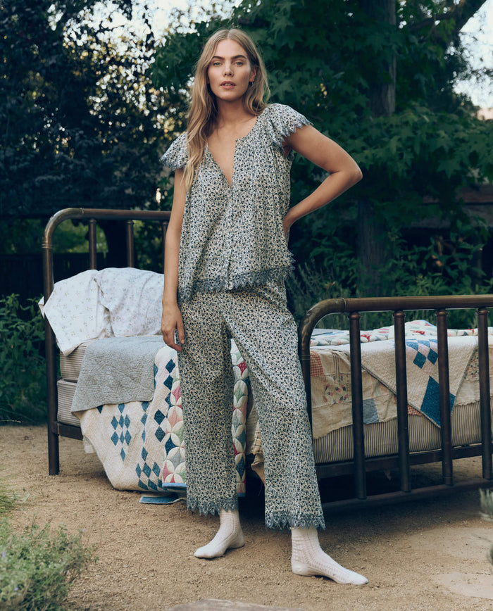 Outdoors by a vintage metal bed topped with patterned quilts, a woman wearing The Great. SP26 Sleep's Eyelet Easy Sleep Pant in Bluebell Ditsy and white socks looks at the camera.