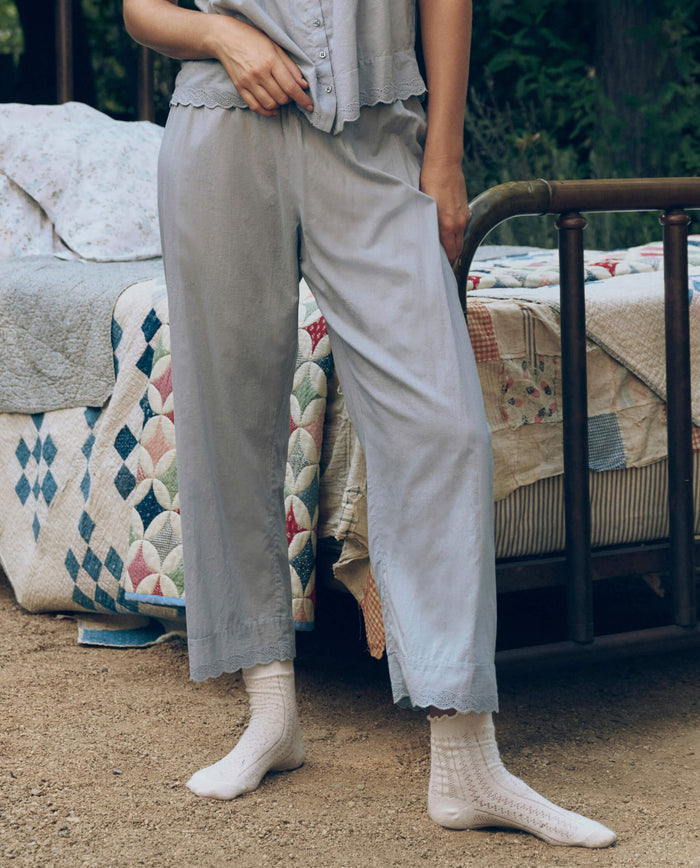 A person in light blue pajamas and white socks stands by a vintage-style bed outdoors, wearing The Great. SP26 Sleep’s The Eyelet Easy Sleep Pant in Rainwater for a delicate, timeless touch.