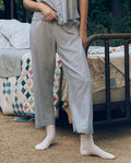 A person in light blue pajamas and white socks stands by a vintage-style bed outdoors, wearing The Great. SP26 Sleep’s The Eyelet Easy Sleep Pant in Rainwater for a delicate, timeless touch.