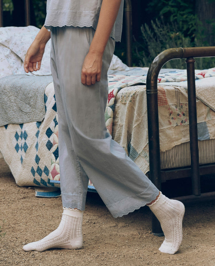 A person wearing The Great. SP26 Sleep The Eyelet Easy Sleep Pant in Rainwater and white socks stands next to a vintage-style bed with a patchwork quilt outdoors.