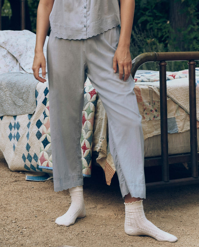 Wearing The Great. SP26 Sleep Eyelet Easy Sleep Pant in Rainwater, a person stands on dirt ground beside a metal-framed bed with a patchwork quilt, showcasing delicate Victorian eyelet lace details for a charming touch.
