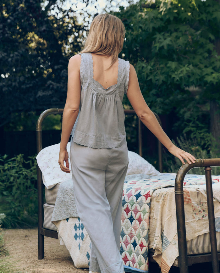 A person in The Great. SP26 Sleep Eyelet Easy Sleep Pant in Rainwater stands outdoors beside a brass bed with patterned quilts, surrounded by greenery.