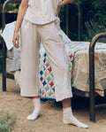 A person stands by a metal-framed bed outdoors, dressed in THE GREAT. CORE SLEEP The Eyelet Easy Sleep Pant in True White with a sleeveless top and white socks; colorful patchwork quilts cover the bed.