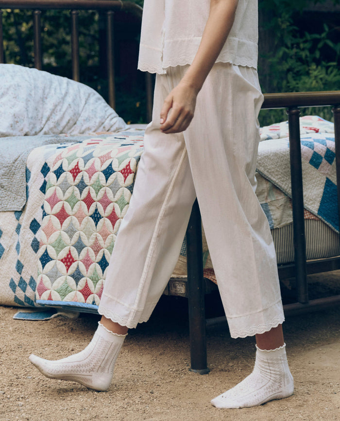 A person in THE GREAT. CORE SLEEP's Eyelet Easy Sleep Pant in True White stands beside a bed with a colorful quilt outdoors, wearing white socks and Victorian-inspired loungewear.