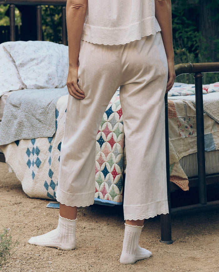 A person in THE GREAT. CORE SLEEP's True White Eyelet Easy Sleep Pant stands by a bed with a patchwork quilt on a dirt surface, their delicate white cotton voile pants and socks enhancing the relaxed, Victorian-inspired scene.