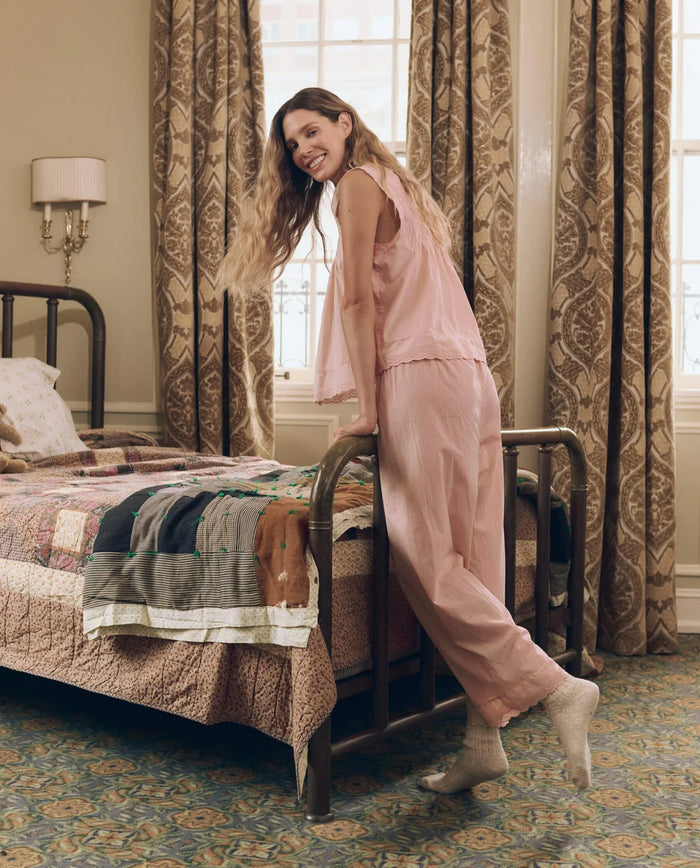 Wearing The Eyelet Easy Sleep Pant in Washed Rose by THE GREAT. F26 SLEEP, a woman leans against the foot of a metal bed in a vintage-inspired bedroom adorned with patterned curtains and a patchwork quilt, creating an effortlessly elevated sense of comfort.