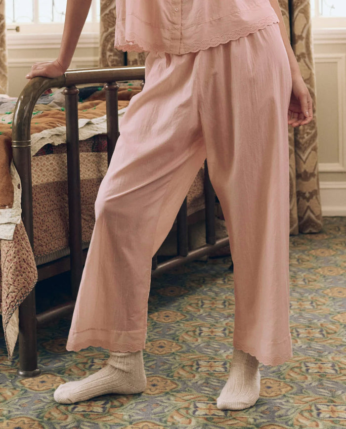 A person stands indoors beside a vintage-style bed with a patchwork quilt on a patterned carpet, radiating elevated comfort in THE GREAT. F26 SLEEP’s Eyelet Easy Sleep Pant in Washed Rose—crafted from airy cotton voile—and cream socks.