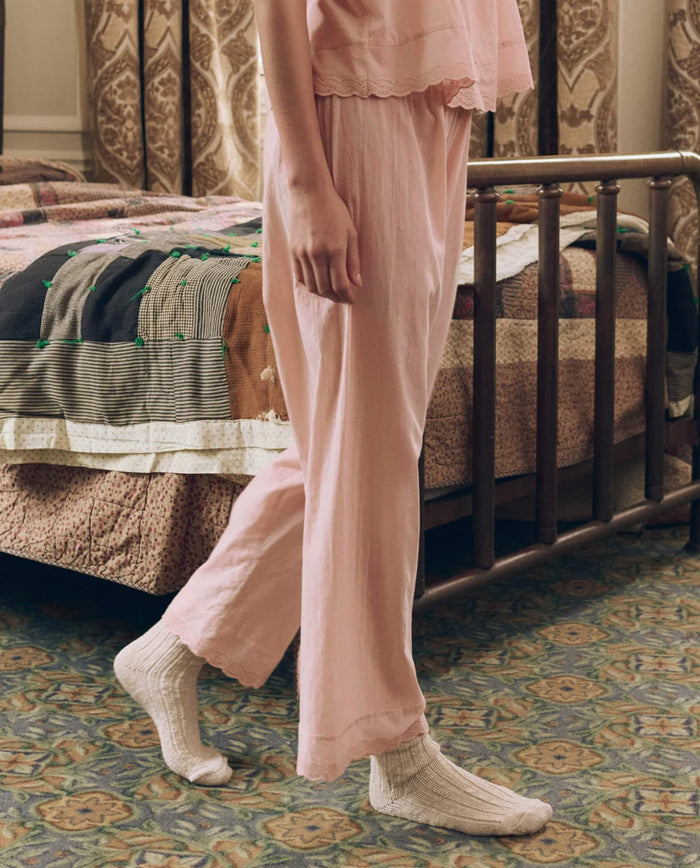 A person in THE GREAT. F26 SLEEP’s The Eyelet Easy Sleep Pant in Washed Rose and white socks stands by a vintage metal bed with a patchwork quilt in a carpeted room featuring patterned curtains, embodying the elevated comfort and cozy style of these soft sleep pants.