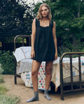 A woman in The Eyelet Mini Tank Dress in Black by The Great. SP26 Sleep and gray socks stands outside by a metal bed with patchwork quilts, surrounded by greenery and trees.