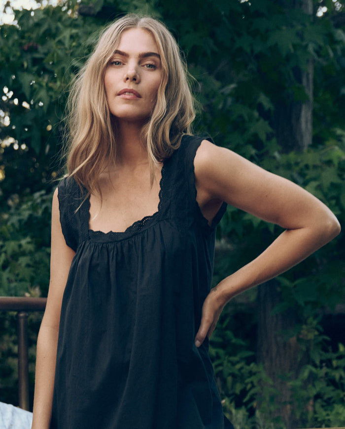 A woman with long, wavy blonde hair stands outdoors in The Great. SP26 Sleep’s black Eyelet Mini Tank Dress, surrounded by lush green foliage.