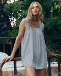 A woman with long blonde hair stands outdoors by a vintage bed, wearing The Great. SP26 Sleep's The Eyelet Mini Tank Dress in Rainwater, with green foliage in the background.