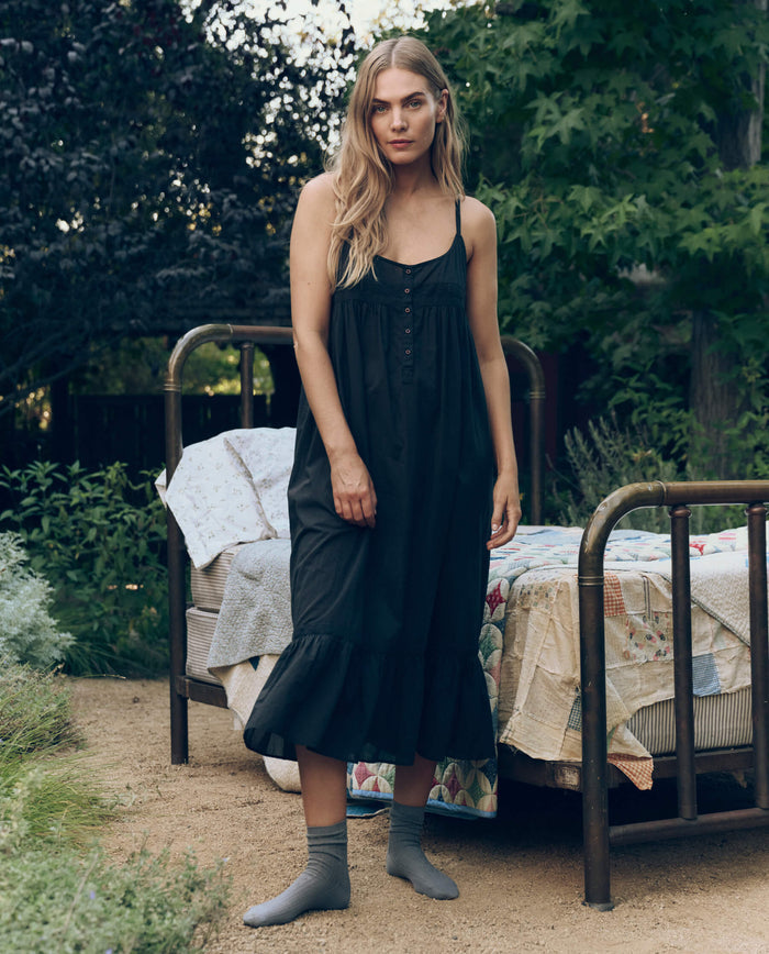 A woman in THE GREAT. CORE SLEEP The Eyelet Ruffle Tank Night Dress in black with gray socks stands outdoors by a vintage bed with patchwork bedding, surrounded by lush trees and plants.