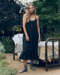 A woman in THE GREAT. CORE SLEEP The Eyelet Ruffle Tank Night Dress in black with gray socks stands outdoors by a vintage bed with patchwork bedding, surrounded by lush trees and plants.