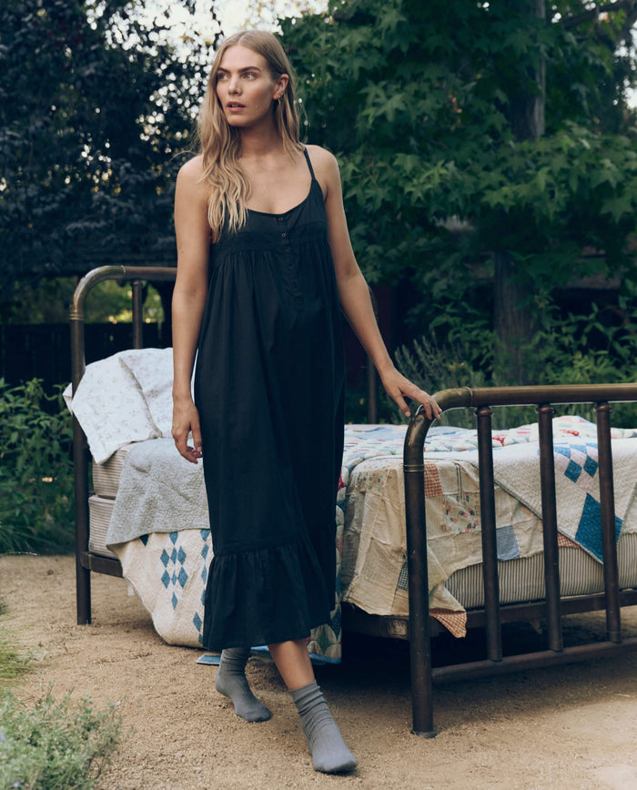 A woman in THE GREAT. CORE SLEEP’s black Eyelet Ruffle Tank Night Dress and gray socks stands by an outdoor bed with patchwork quilts, surrounded by greenery.