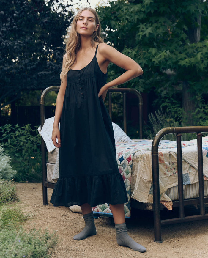 A woman in THE GREAT. CORE SLEEP's black Eyelet Ruffle Tank Night Dress and gray socks stands on dirt beside a metal bed with a patchwork quilt, surrounded by greenery.