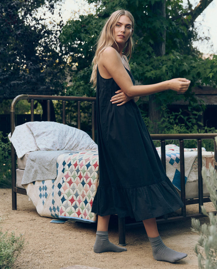 A woman in THE GREAT. CORE SLEEP The Eyelet Ruffle Tank Night Dress in black stands beside an outdoor bed with patterned quilts and pillows, surrounded by greenery.
