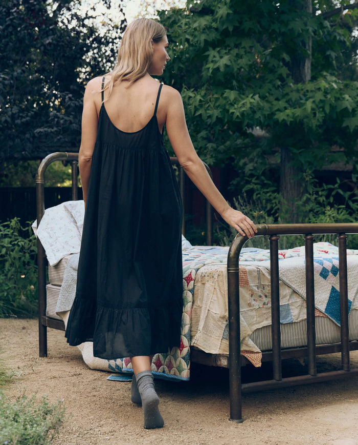 A woman in THE GREAT. CORE SLEEP The Eyelet Ruffle Tank Night Dress in black and gray socks stands outdoors by a vintage metal bed with quilted blankets, surrounded by greenery.