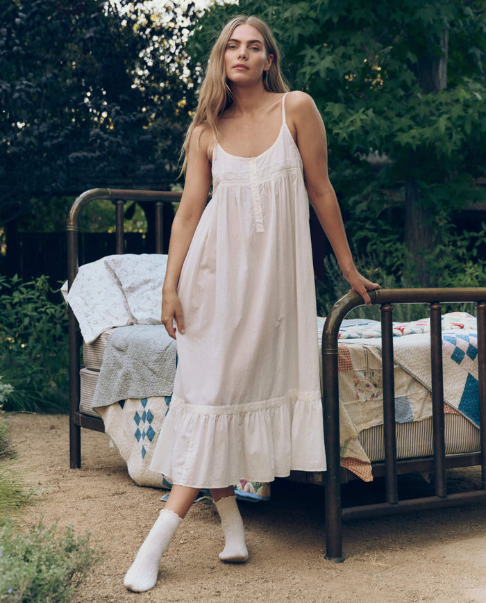 A woman in THE GREAT. CORE SLEEP The Eyelet Ruffle Tank Night Dress in True White stands beside a vintage metal bed with patchwork quilts, surrounded by greenery.