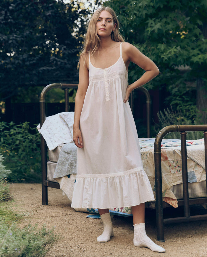 Wearing The Eyelet Ruffle Tank Night Dress in True White by THE GREAT. CORE SLEEP, a woman stands in socks by an outdoor bed with quilts, amid lush greenery and trees, evoking Victorian-inspired lace charm.