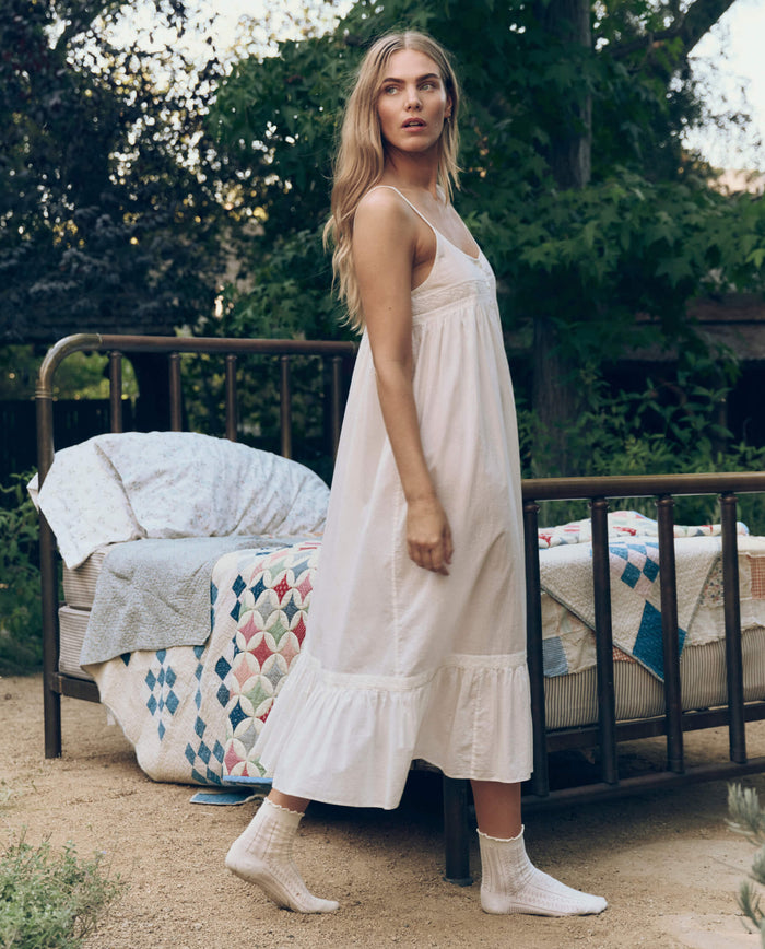 A woman in THE GREAT. CORE SLEEP The Eyelet Ruffle Tank Night Dress in True White and socks stands beside an outdoor metal bed with patterned quilts, surrounded by trees and greenery.