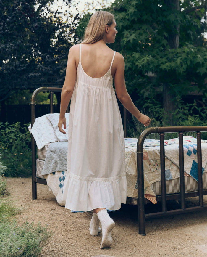 A woman in THE GREAT. CORE SLEEP The Eyelet Ruffle Tank Night Dress in True White and socks walks toward a vintage metal bed with a patchwork quilt, outdoors in a garden.
