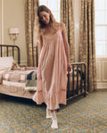 A woman wearing THE GREAT. F26 SLEEP’s Eyelet Ruffle Tank Night Dress in Washed Rose and white socks stands in a vintage-style bedroom with a quilted bed and patterned curtains.
