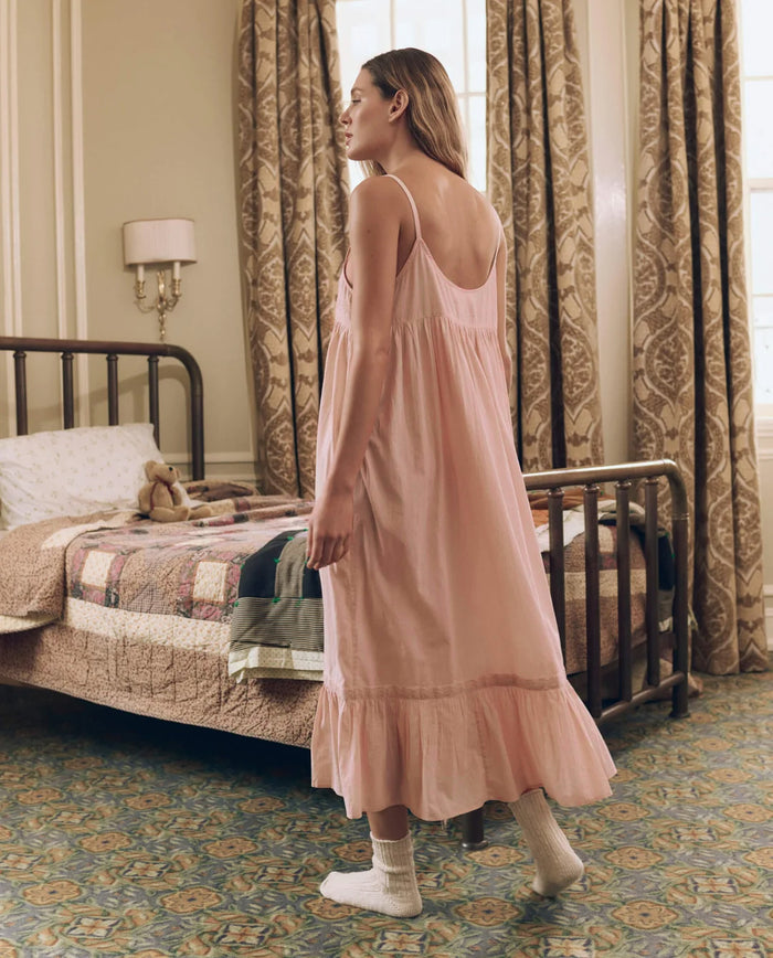 A woman wearing THE GREAT. F26 SLEEP’s Eyelet Ruffle Tank Night Dress in Washed Rose, paired with socks, stands beside a vintage-style bed adorned with a quilt and teddy bear, in a room decorated with patterned curtains and carpet.
