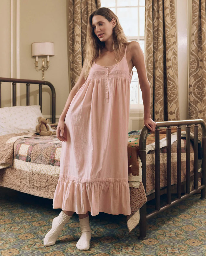 A woman in THE GREAT. F26 SLEEP’s Eyelet Ruffle Tank Night Dress in Washed Rose and white socks stands by a metal bed in a vintage-style bedroom with patterned curtains and a patchwork quilt.