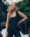 A woman with long blonde hair stands outdoors in front of greenery, wearing THE GREAT. CORE SLEEP The Eyelet Tank in black and matching pants, with one hand on her hip.
