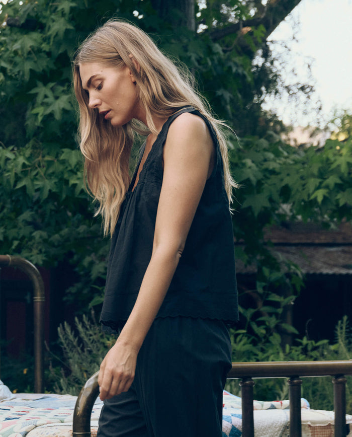 A woman with long blonde hair stands outdoors amidst greenery, wearing THE GREAT. CORE SLEEP’s The Eyelet Tank in black and black pants, beside a brass bed frame draped with a quilt.