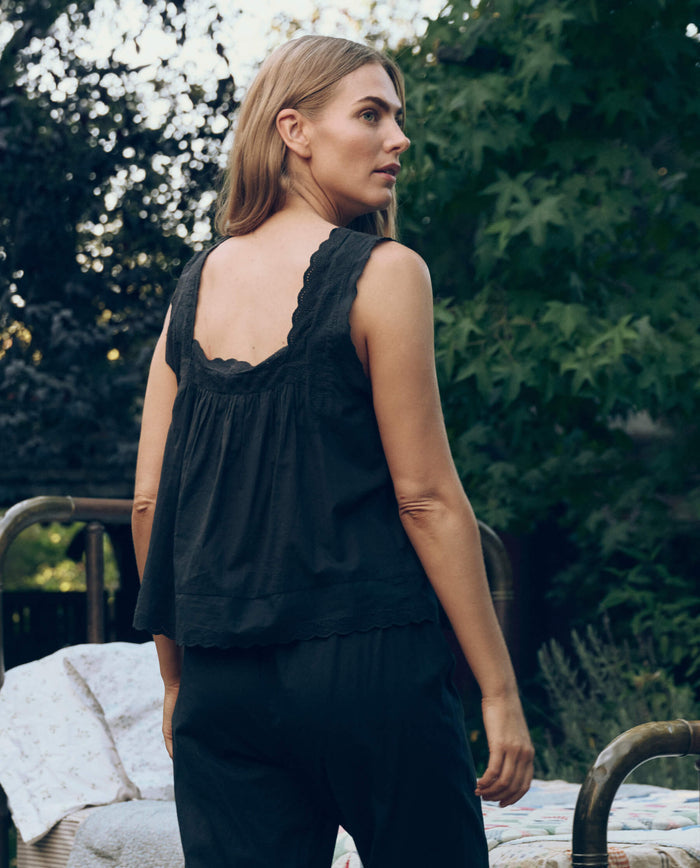 A woman with long hair stands outdoors, wearing THE GREAT. CORE SLEEP's The Eyelet Tank in black and pants, looking over her shoulder with trees and bedding visible in the background.