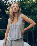 A woman with long blonde hair stands outdoors wearing The Great. SP26 Sleep's The Eyelet Tank in Rainwater, with greenery and a bed frame in the background.