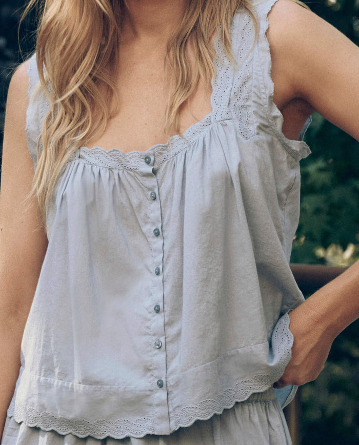 A woman outdoors wears The Great. SP26 Sleep's Eyelet Tank in Rainwater, with lace trim, buttons, and airy cotton voile fabric, her hand in her pocket showing off the tank’s flowy silhouette.