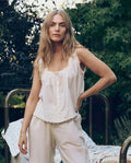 A woman with long blonde hair stands outdoors in a sleeveless The Eyelet Tank in True White by THE GREAT. CORE SLEEP, paired with matching pants, surrounded by greenery and an outdoor bed.