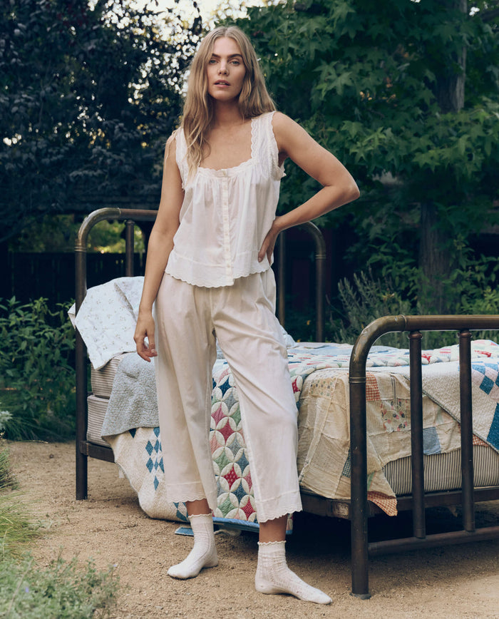 A woman in THE GREAT. CORE SLEEP The Eyelet Tank in True White and socks stands by a vintage metal bed draped in patchwork quilts outdoors, surrounded by greenery.