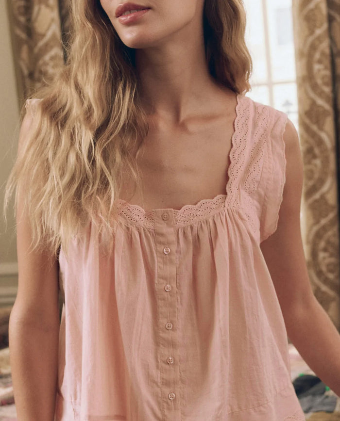 A woman wearing The Eyelet Tank in Washed Rose by THE GREAT. F26 SLEEP—a sleeveless, light pink button-up top with lace trim and a flowy silhouette—stands indoors in front of patterned curtains and a window.