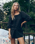 A woman with long blonde hair wears THE GREAT. CORE SLEEP’s The Eyelet Tap Short in black, standing outdoors near a bed with a patterned quilt, surrounded by trees and greenery.