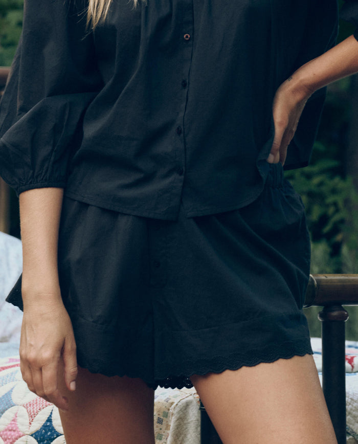 Someone stands outdoors with one hand on their hip, wearing THE GREAT. CORE SLEEP's The Eyelet Tap Short in black and a matching button-up shirt.