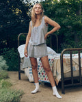 A woman stands outdoors in greenery beside a metal-framed bed with a patchwork quilt, wearing The Eyelet Tap Short -- Rainwater by The Great. SP26 Sleep and white socks.