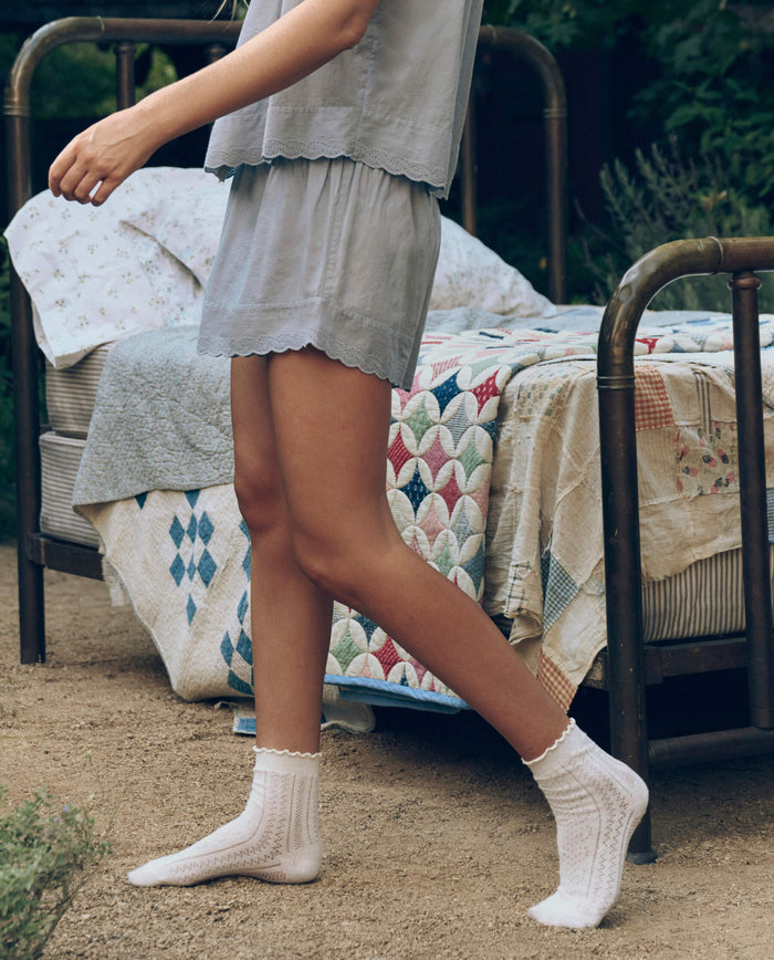 Wearing The Great. SP26 Sleep's Eyelet Tap Short in Rainwater, someone walks barefoot on dirt next to a metal bed with a patchwork quilt outdoors, paired with white socks.