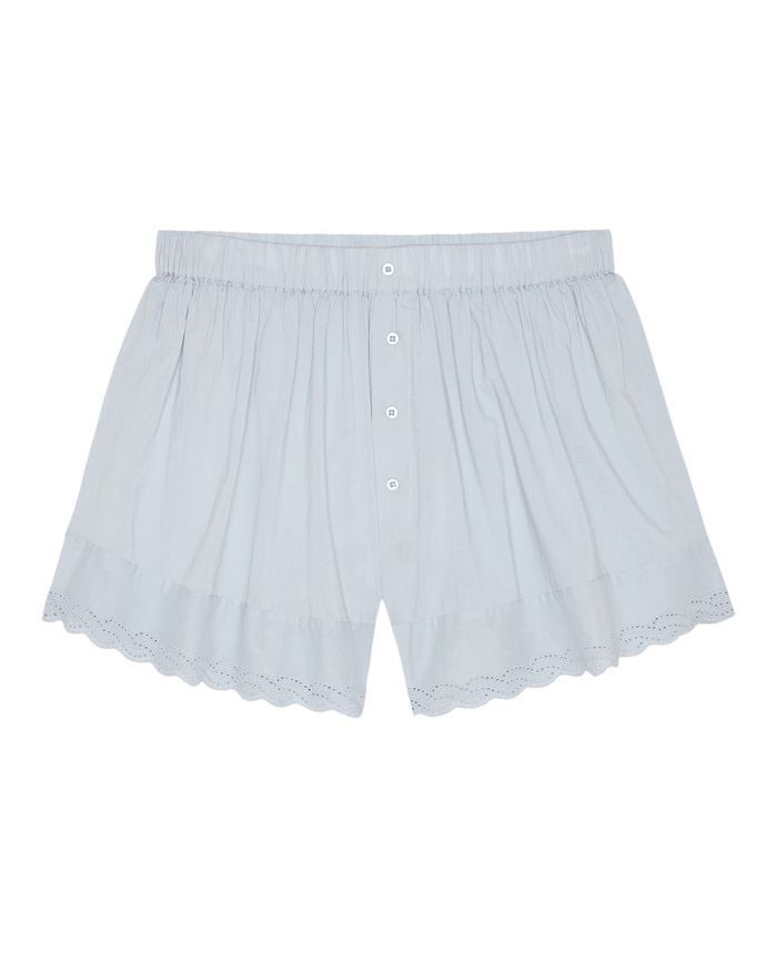 The Great. SP26 Sleep Eyelet Tap Short in Rainwater: light blue cotton voile sleep short with elastic waistband, front button closure, scalloped hem, and eyelet lace trim along the bottom edges, shown on a white background.